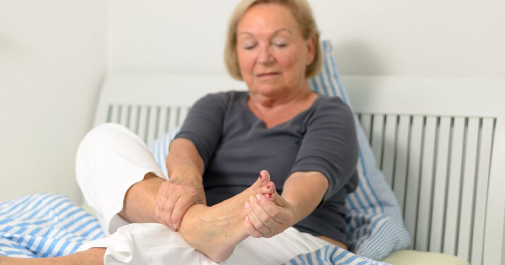 Older woman sitting on bed holding her foot with concerned expression, experiencing numbness and tingling from peripheral neuropathy
