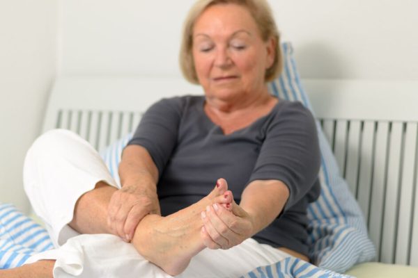 Older woman sitting on bed holding her foot with concerned expression, experiencing numbness and tingling from peripheral neuropathy